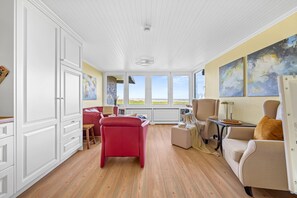 Une pièce avec une grande fenêtre, un fauteuil rouge, un canapé beige et une petite table sur laquelle se trouvent un livre et une assiette de noix.