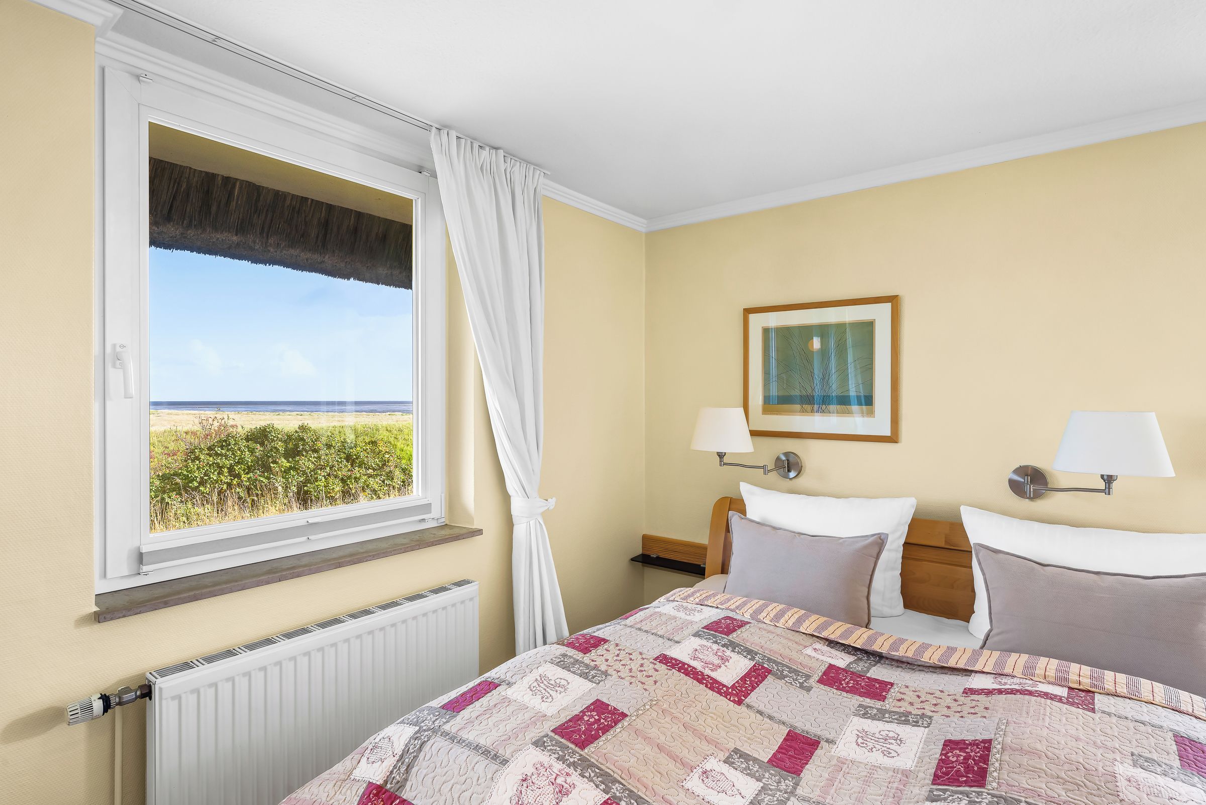 Ein Hotelzimmer mit Bett, Fenster mit Blick auf den Strand, Nachttisch mit Lampe und einem gerahmten Bild an der Wand.