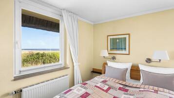 Ein Hotelzimmer mit Bett, Fenster mit Blick auf den Strand, Nachttisch mit Lampe und einem gerahmten Bild an der Wand.