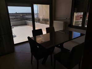 A modern kitchen with a glass dining table and chairs, a view of a balcony with a railing, and a radiator on the wall. - Sky Line Penthouse Apartment (Trikala)