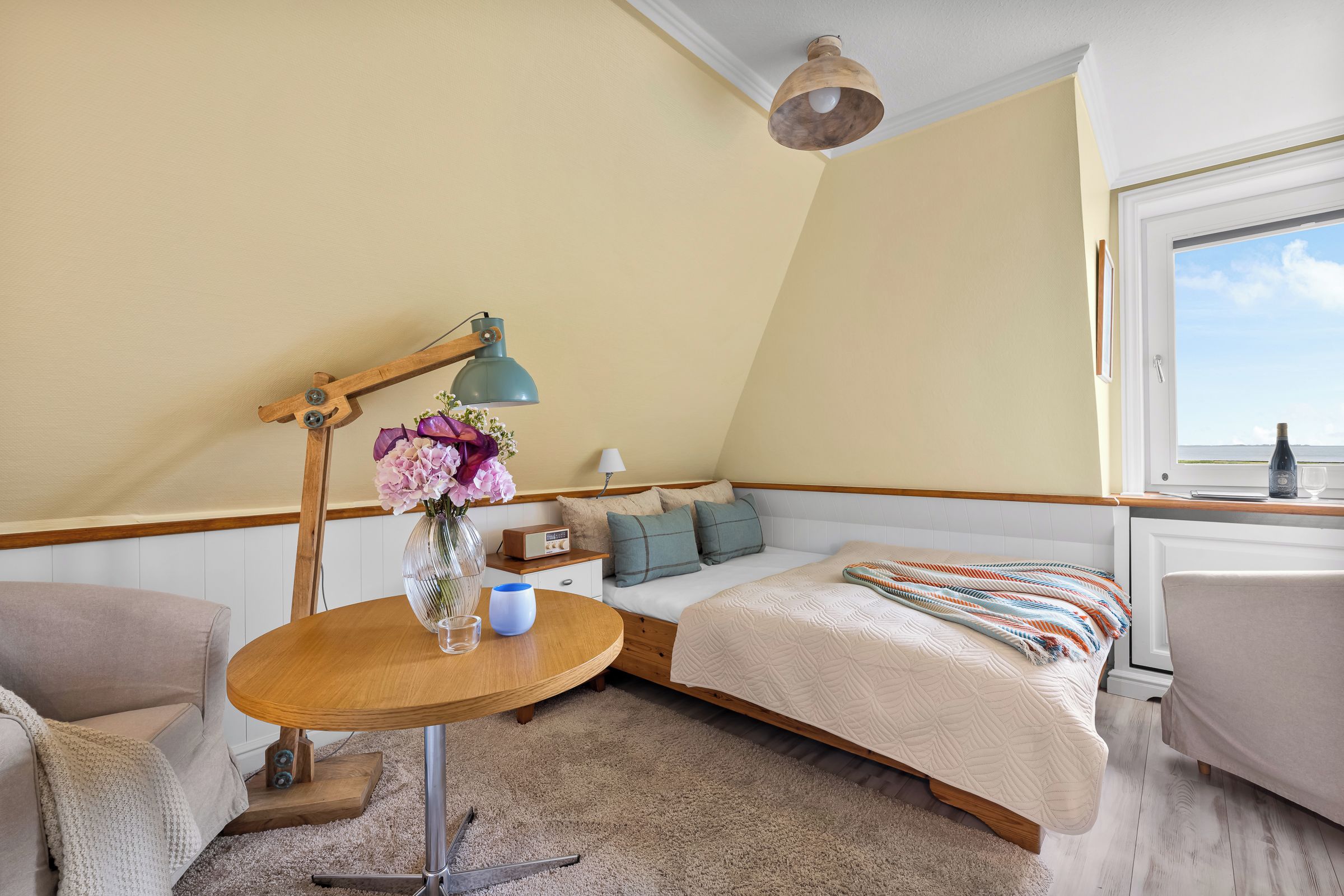 A bedroom with a bed, a table with a lamp, a vase with flowers, and a window with a view.