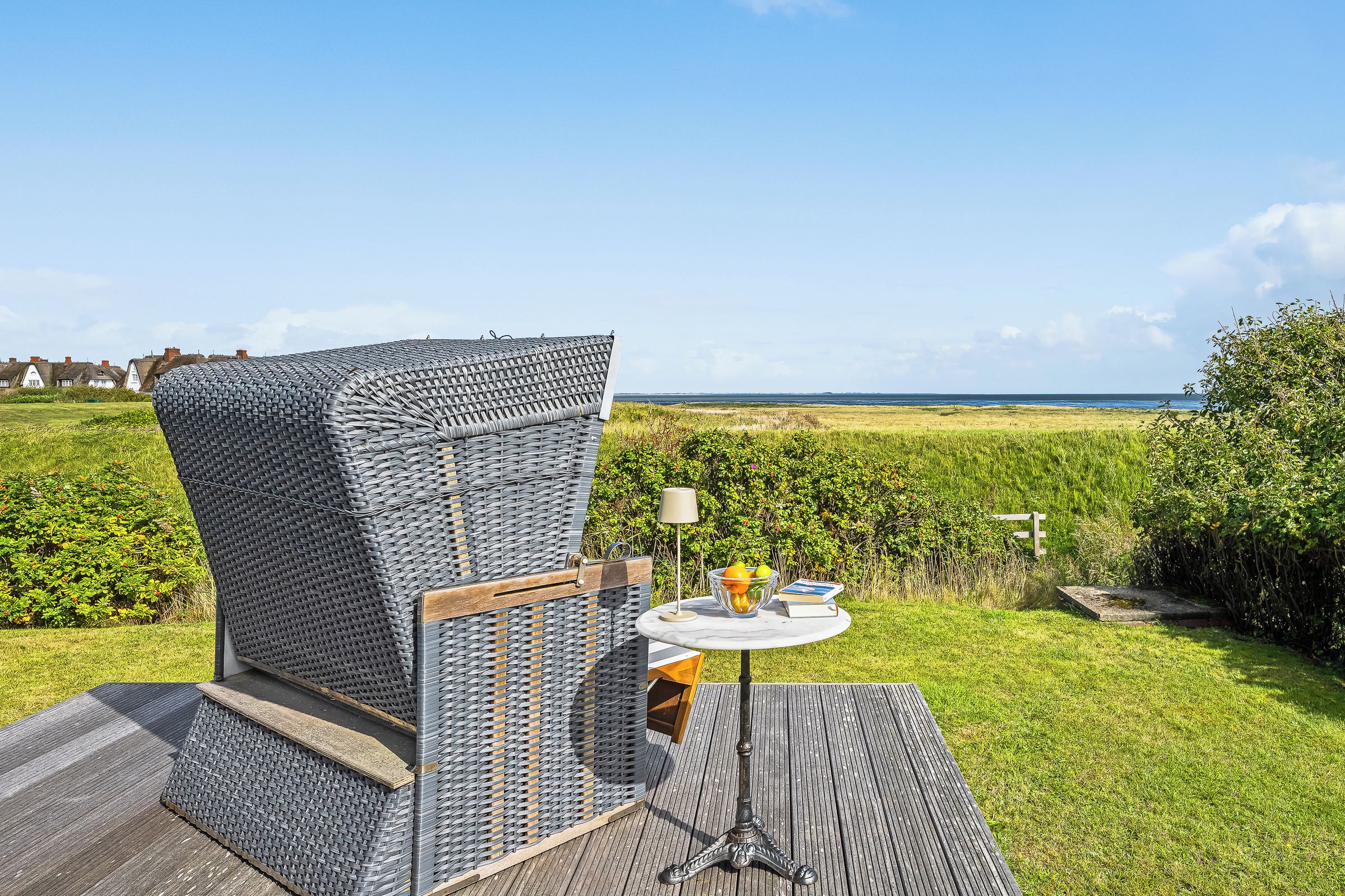 A wicker chair with a view of the sea, a round table with a lamp, and a book.