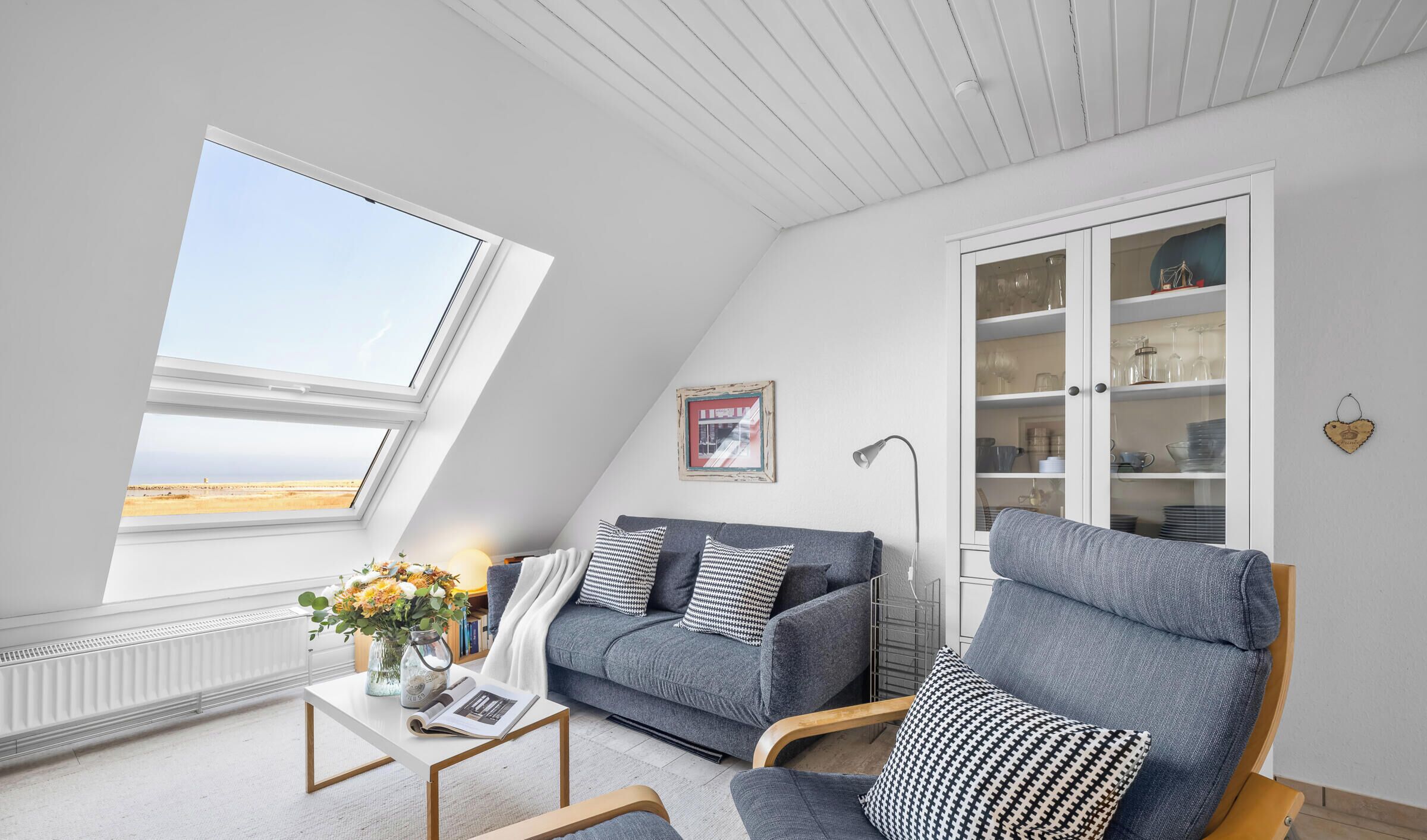 A room with a sloped ceiling, a sofa, a chair, a cabinet with glass doors, and a view of the beach.