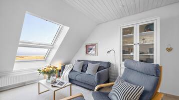 A room with a sloped ceiling, a sofa, a chair, a cabinet with glass doors, and a view of the beach.