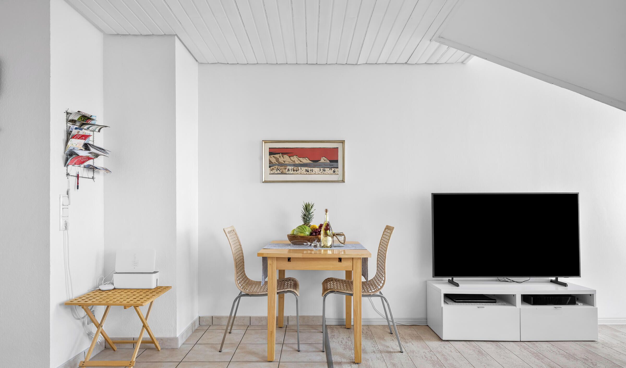 A modern living room with a dining table, two wicker chairs, a TV on a white cabinet, a framed picture on the wall, and a small shelf with magazines.