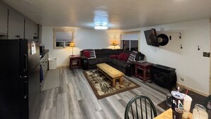 Un espace de vie compact comprenant un coin cuisine, un ensemble de canapés, une table basse et un téléviseur à écran plat fixé au mur.