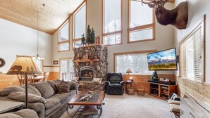 A cozy living room with a stone fireplace, large windows, a mounted deer head, and a television. - Paradise in the Pines | Hot Tub | 35 miles to YNP (Island Park)