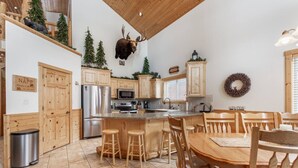 A kitchen with wooden cabinets, a stainless steel refrigerator, a microwave, and a wooden dining table with chairs. - Paradise in the Pines | Hot Tub | 35 miles to YNP (Island Park)