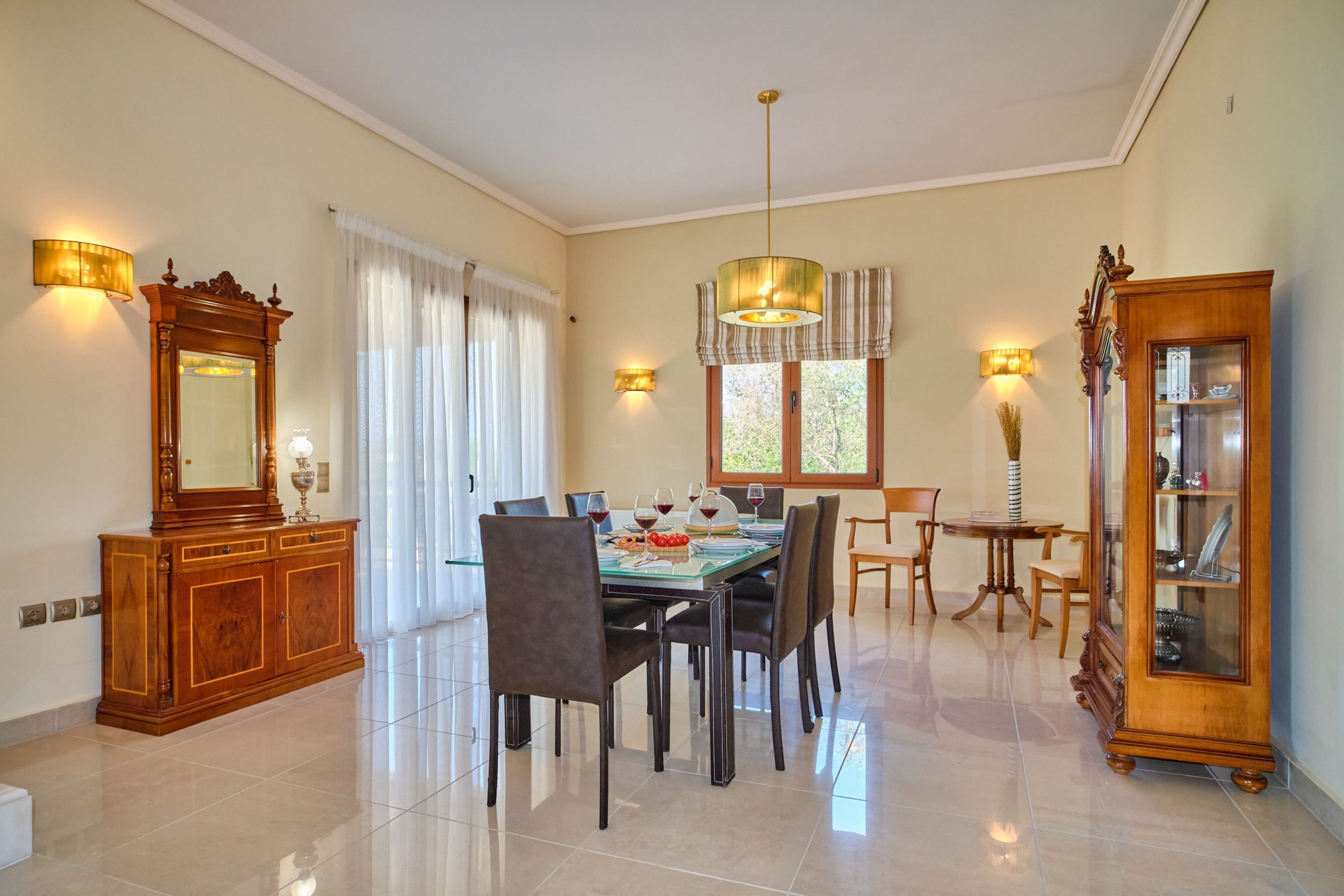 Un comedor con una mesa dispuesta para cuatro personas, rodeada de sillas, una araña y un gran aparador de madera con puertas de cristal.