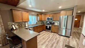A modern kitchen with wooden cabinets, stainless steel appliances, a large island with bar stools, and a view of the outdoors through the window. - Cozyville Cabin: Quiet Getaway, Built 2019 (Big Bear)