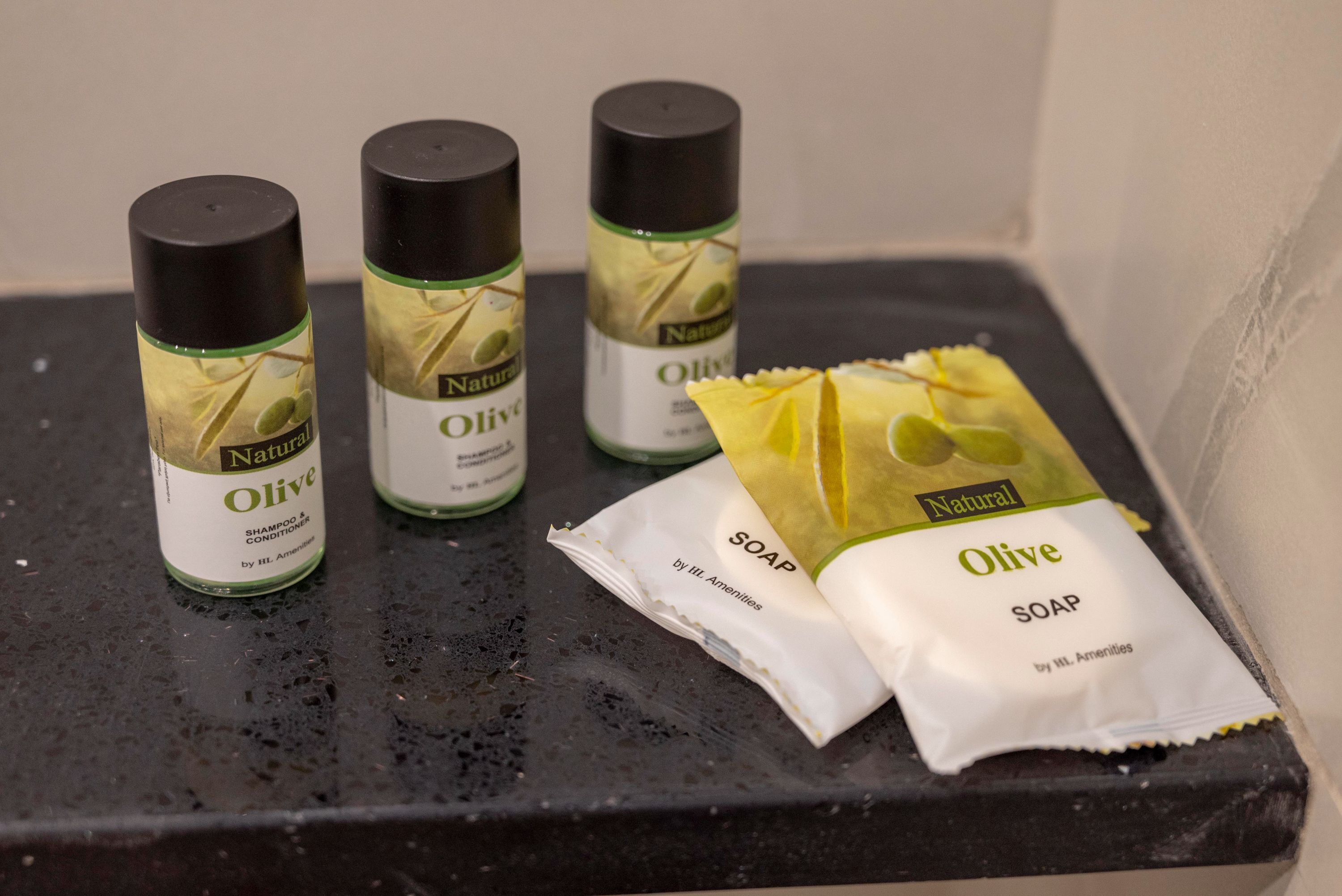 Trois bouteilles de shampoing et d’après-shampoing à base d’huile d’olive naturelle, ainsi qu’un paquet de savon à l’olive, sont posés sur une surface noire.