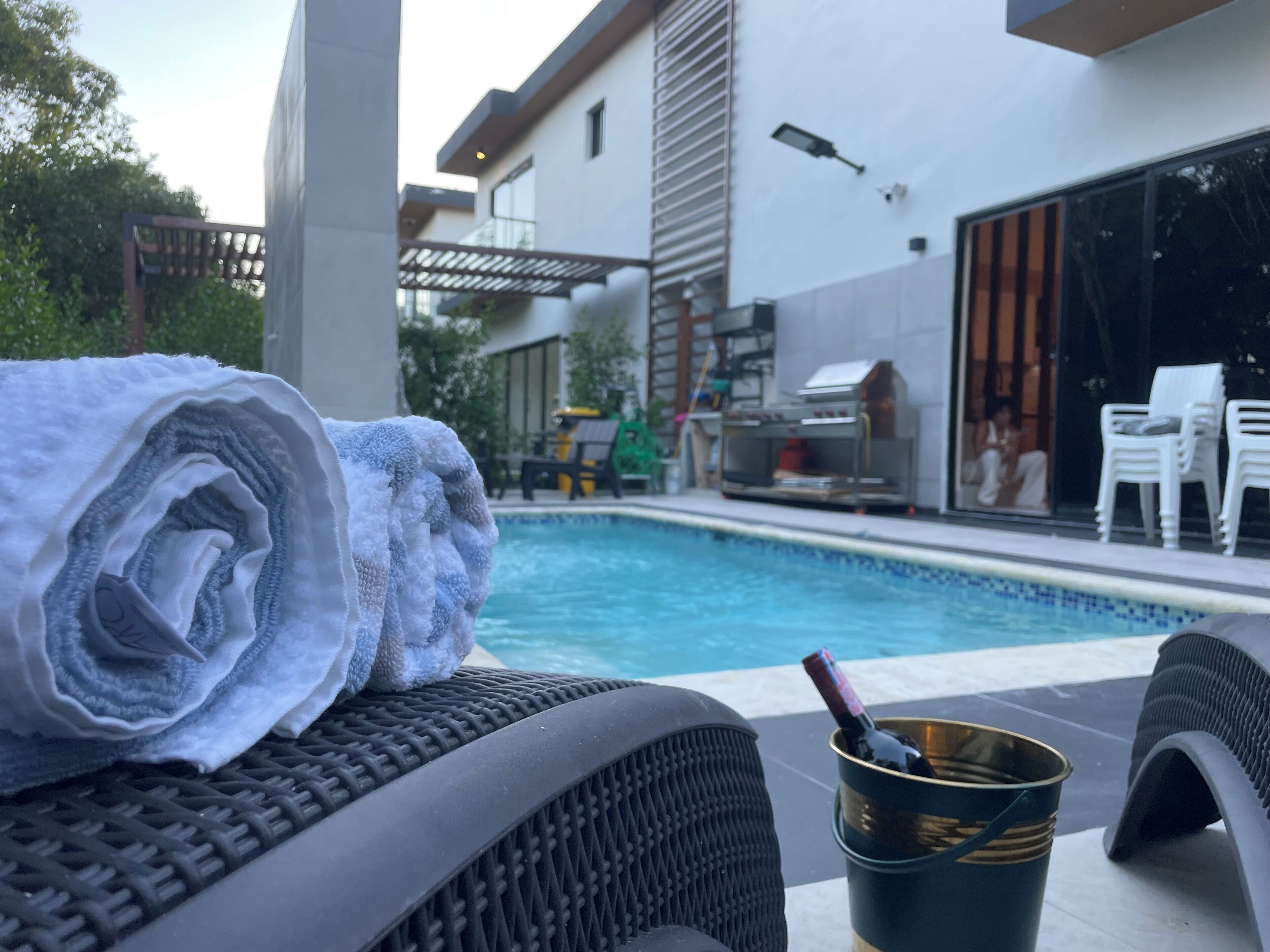 A poolside area with a lounge chair, a rolled towel, and a bucket with a bottle.