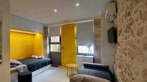 A compact bedroom with a bed, a small seating area, and a window with blinds. - [Bracket] in Heraklion (Heraklion)