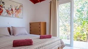 Ein Schlafzimmer mit einem Bett, einer Holzkommode und einem großen Fenster mit Blick auf Grünflächen.