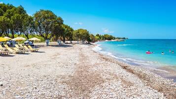 Una playa de guijarros con tumbonas y sombrillas, un mar azul cristalino y árboles a lo largo de la orilla.