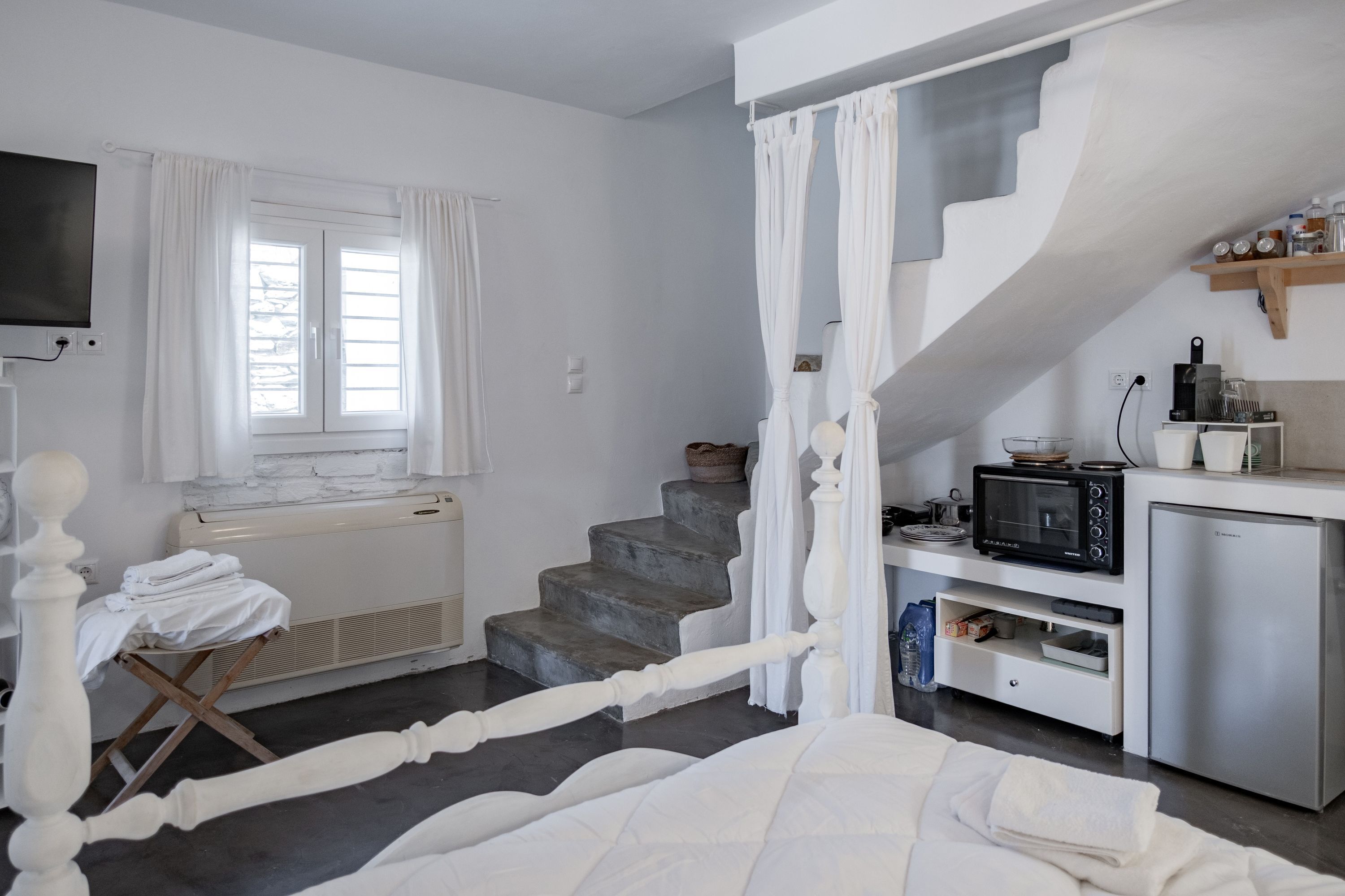 Een slaapkamer met een bed, een klein tafeltje met een stoel, een raam met gordijnen, een trap en een kitchenette met een magnetron en een koelkast.