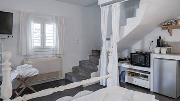 Een slaapkamer met een bed, een klein tafeltje met een stoel, een raam met gordijnen, een trap en een kitchenette met een magnetron en een koelkast.