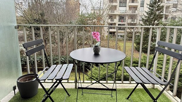Un balcon avec un revêtement de gazon synthétique, une table ronde, deux chaises pliantes et un pot de fleurs.