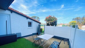 A patio with a white table and two blue chairs, a white building with a window, and a tree with pink flowers. - Cosy, quiet house close to the oyster port. (La Teste-de-Buch)