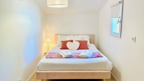 A neatly made bed with a heart-shaped pillow, a bedside lamp, and a window with blinds. - Cosy, quiet house close to the oyster port. (La Teste-de-Buch)