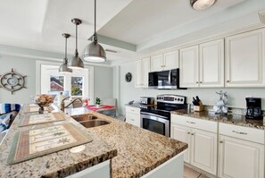 A kitchen with granite countertops, white cabinets, stainless steel appliances, pendant lighting, and a visible window. - Tybee Dream: 2 Bed 2 Bath Condo w/ rooftop Pool (Tybee Island)