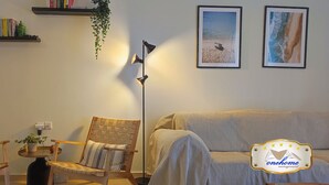 A cozy living room with a sofa, two wooden chairs, a floor lamp, and wall-mounted shelves with books and a hanging plant. - Filoxenia  in the centre of Kalamata (Kalamata)