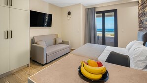 A hotel room with a bed, a sofa, a dining table with fruit, a TV, and a view of the beach. - Grande Mare with Sea view (Gazi)