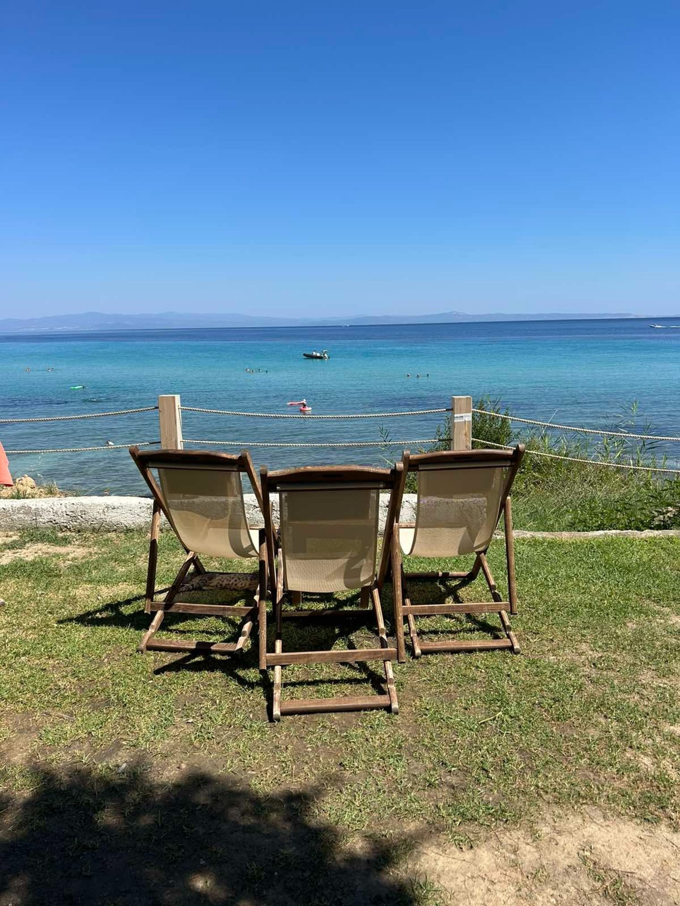 Three wooden lounge chairs facing a clear blue sea with a distant view of the coastline.