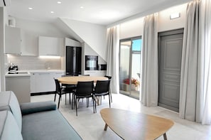 A modern kitchen with white cabinets, a dining area with a wooden table and black chairs, and a sliding glass door leading to a balcony with curtains. - Suite Christina - Demar Suites (Vlicho)