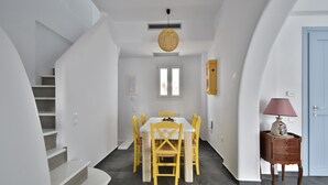 Un coin repas avec une table blanche et des chaises jaunes, un escalier à gauche, une porte bleue à droite et une petite table d’appoint en bois avec une lampe.