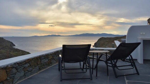 Une terrasse avec deux chaises et une table, d’où l’on a une vue sur la mer, avec un mur de pierre et un bâtiment blanc en arrière-plan.