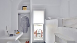 A compact, white-themed room with a laptop on a small table, a hanging coat, and a view of an outdoor seating area through a glass door. - Tinos Portraits Ano Meria (Exomvourgo)
