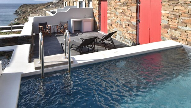 A pool area with a stone wall, red doors, and a white building with a pergola. - Tinos Portraits Ano Meria (Exomvourgo)
