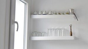 A white kitchen with a window, a sink, and shelves with glassware and a tea pot. - Tinos Portraits Ano Meria (Exomvourgo)