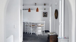 A narrow hallway with a white table and chairs, a wooden chest, and a white radiator. - Tinos Portraits Kato Meria (Exomvourgo)