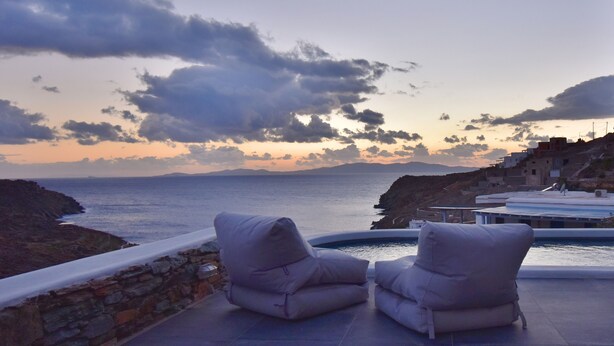 A terrace with two cushioned chairs overlooking a body of water and a mountainous landscape. - Tinos Portraits Kato Meria (Exomvourgo)