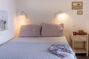 A bedroom with a bed, two bedside lamps, a nightstand with a candle and a hat, and a wall with a painting.