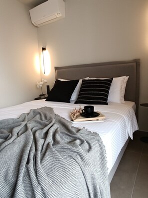 Una cama bien hecha con un edredón gris, una mesita de noche con una lámpara y una bandeja con una taza y un objeto decorativo.