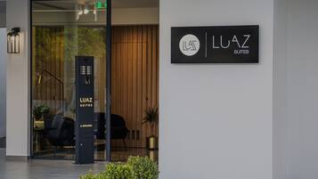 Een moderne gebouw-ingang met een glazen deur, een zwarte kiosk met de tekst 'LUAZ SUITES' en een bord met dezelfde naam.