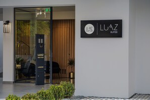 Een moderne gebouw-ingang met een glazen deur, een zwarte kiosk met de tekst 'LUAZ SUITES' en een bord met dezelfde naam.