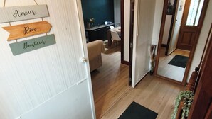 En hall med trägolv, en svart matta och en skylt med text på franska. Därifrån leder en dörr in till ett rum med en säng och ett skrivbord.