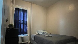 Una stanza piccola e poco illuminata, con un letto singolo, un termosifone e una finestra con tende.