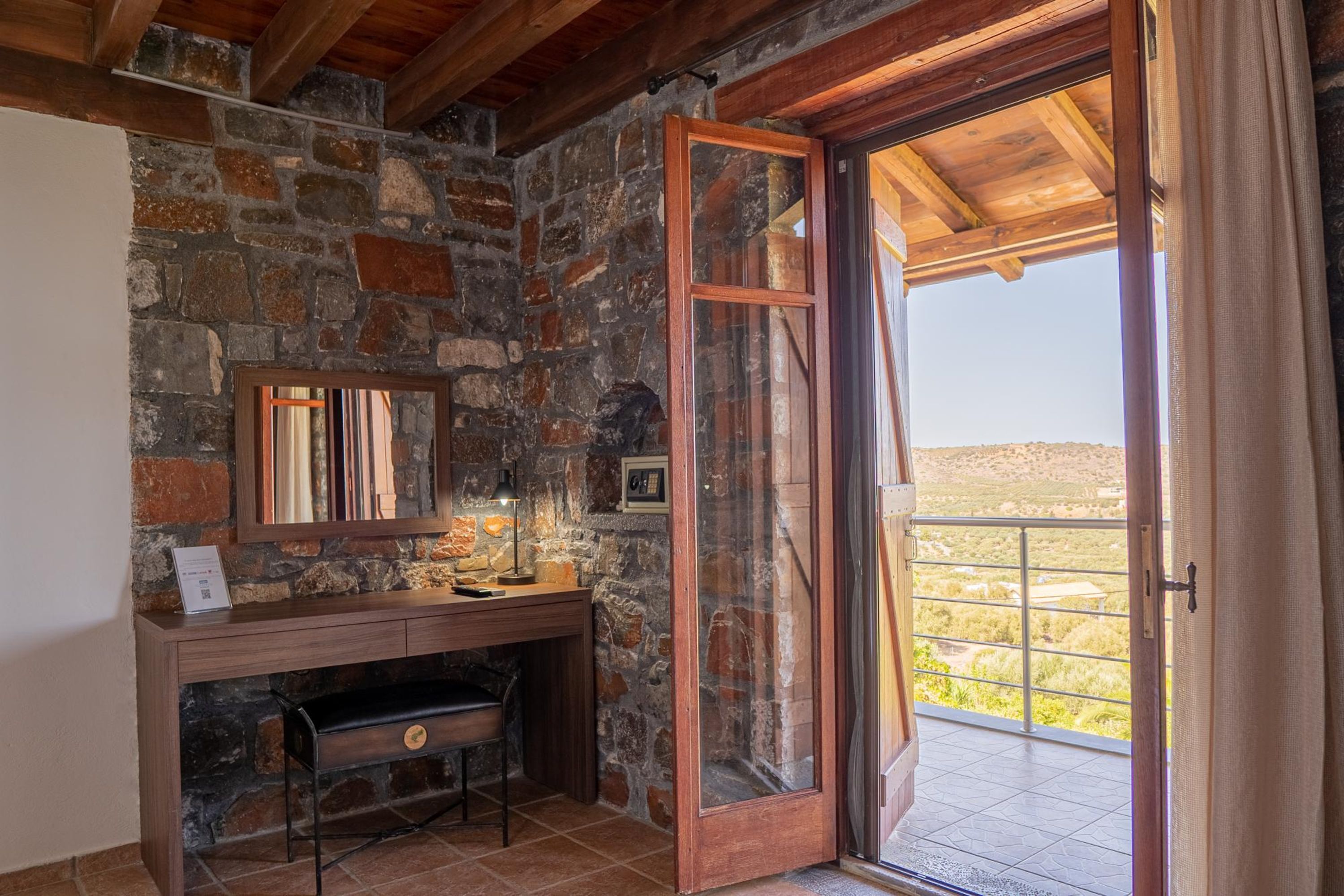 Een betegelde badkamer met een houten wastafel, een spiegel en een deur die leidt naar een balkon met uitzicht op het landschap.