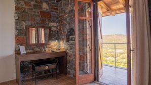 Ein Badezimmer mit Steinsäulen, einem Holzkosmetikschrank, einem Spiegel und einer Tür, die auf einen Balkon mit Blick auf die Landschaft führt.