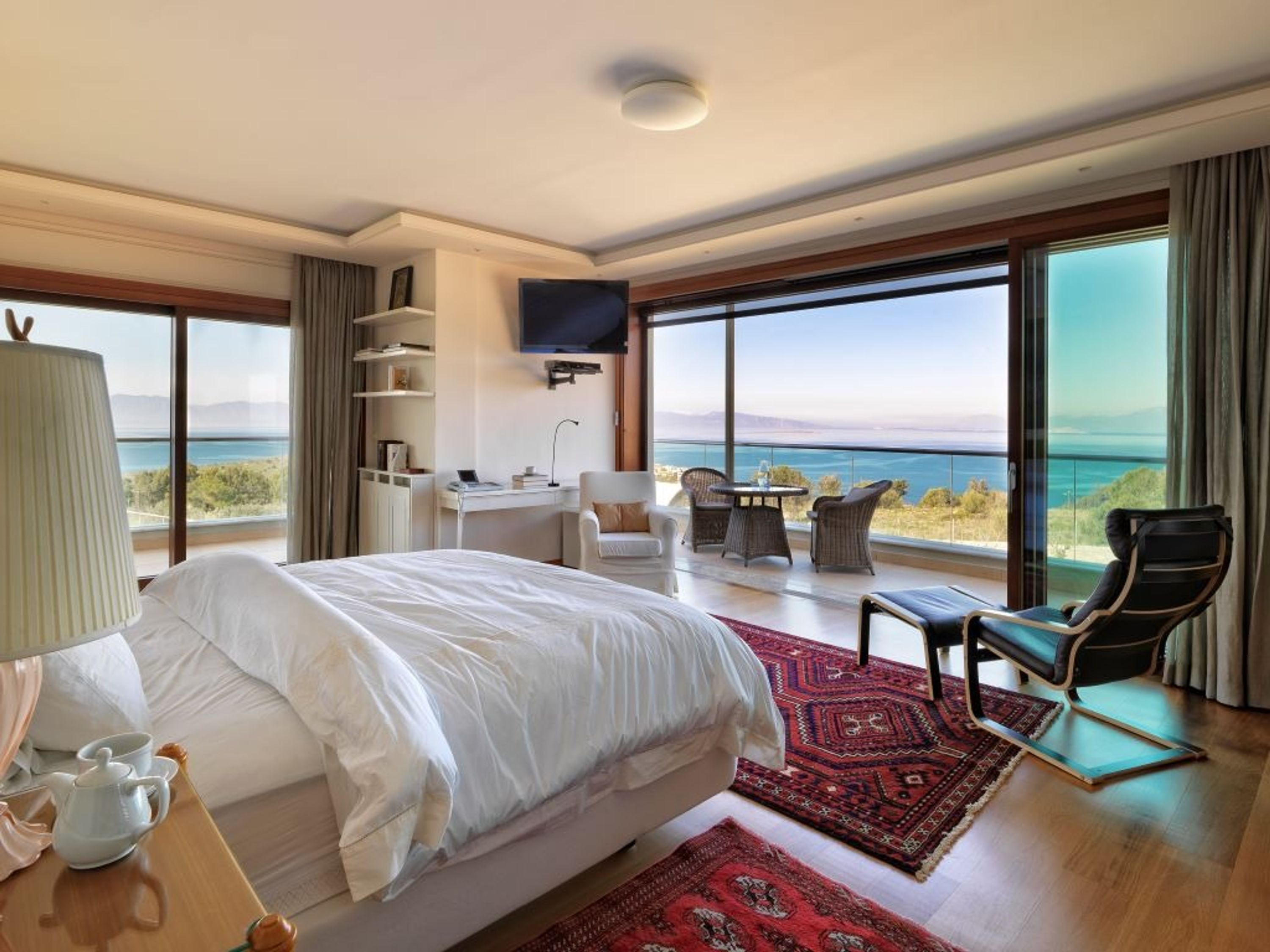 A spacious bedroom with a large bed, a desk, and a chair, all facing a balcony with a view of the sea.