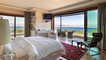 A spacious bedroom with a large bed, a desk, and a chair, all facing a balcony with a view of the sea.