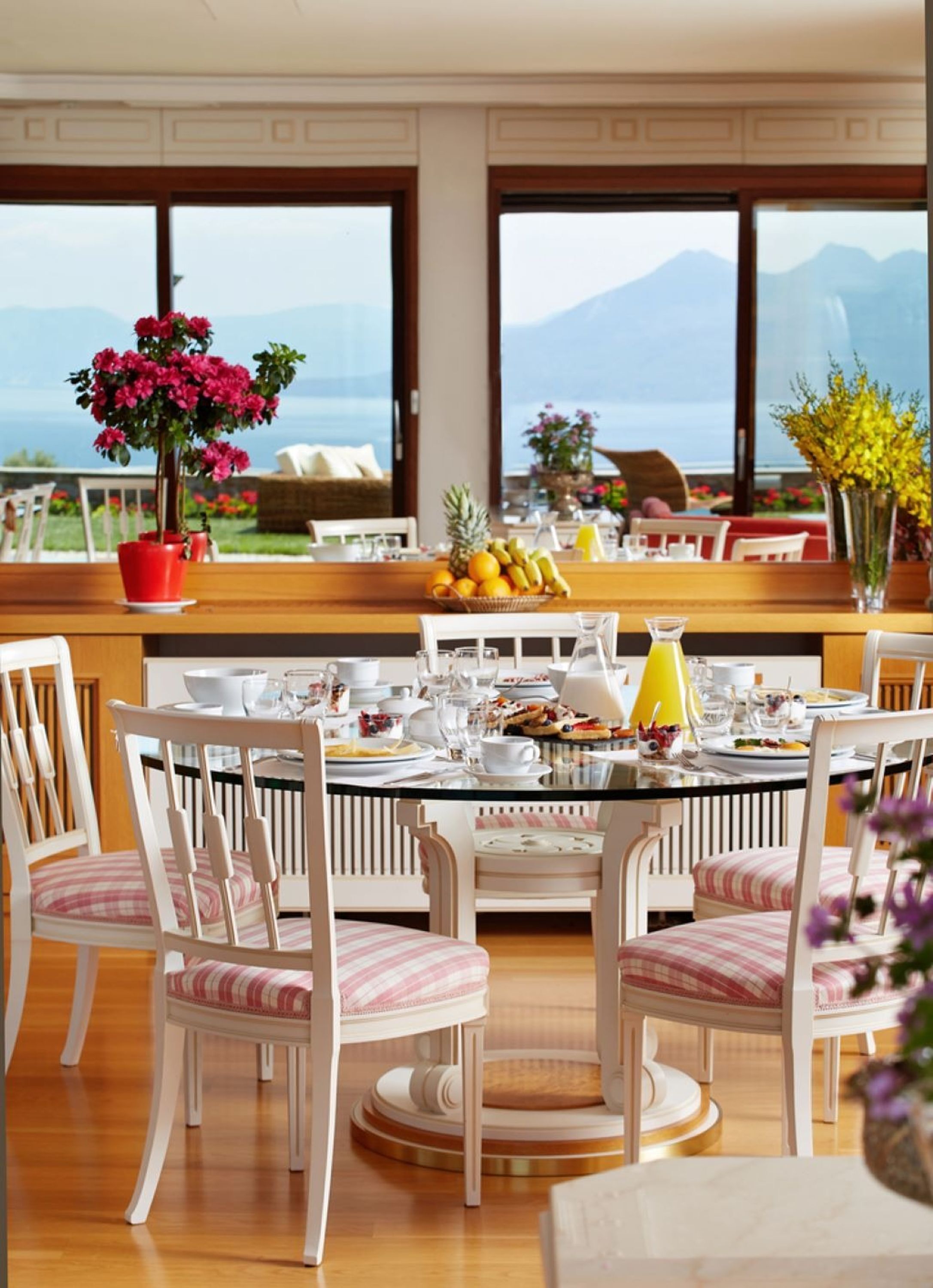 A dining area with a round table set for four, surrounded by white chairs with striped cushions. A potted plant with pink flowers is on the left side of the table.