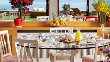 A dining area with a round table set for four, surrounded by white chairs with striped cushions. A potted plant with pink flowers is on the left side of the table.