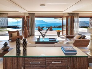 A spacious living room with a glass coffee table, comfortable seating, and a view of the ocean and mountains. - Armonia Luxury Villa (Perdika)