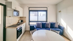 A compact kitchen with a microwave, stove, and sink. A blue sofa with cushions is positioned near a window, and a white table is in the foreground. - Ben Yehouda Obsidian (Tel Aviv-Yafo)
